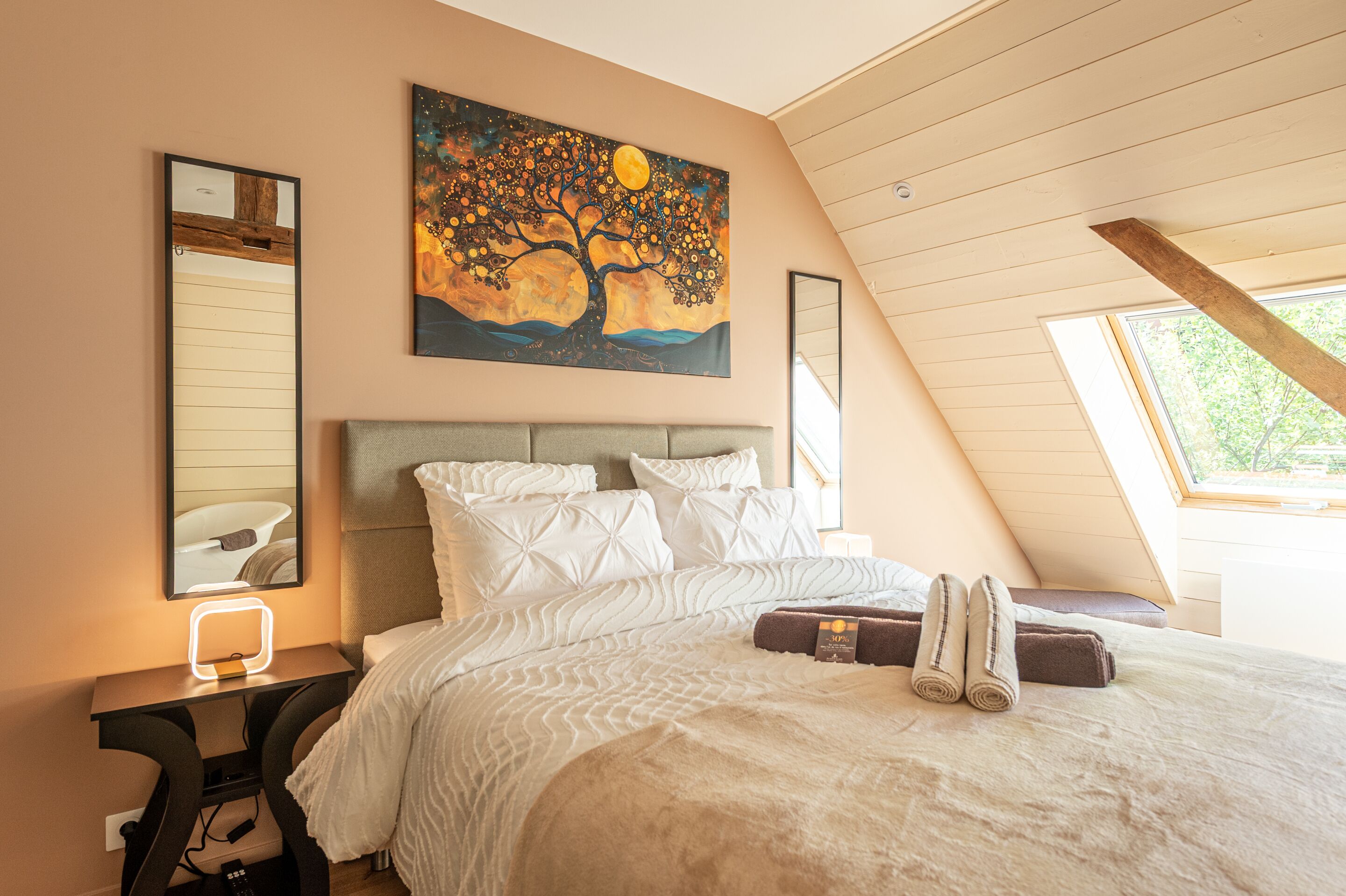 A bedroom with a large bed, a nightstand with a lamp, a painting on the wall, and a window with a view.