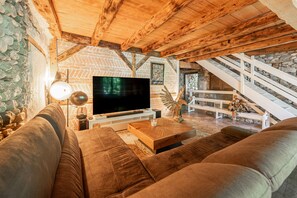 A cozy living room with a large flat-screen TV, comfortable sofas, a wooden coffee table, and a stone wall.
