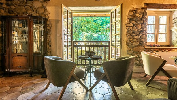 A room with a stone wall, a glass cabinet, a table with chairs, and a view of greenery outside. - Le Moulin de mes garçons (Alby-sur-Chéran)