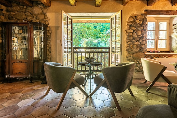 A room with a stone wall, a glass cabinet, a table with chairs, and a view of greenery outside.