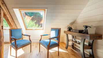 A room with wooden floors, two blue upholstered chairs, a wooden table with a kettle, and a window with a view of greenery.