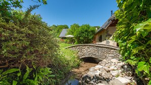 A stone bridge over a stream, surrounded by greenery and a house with a sloped roof. - Le Moulin de mes garçons (Alby-sur-Chéran)