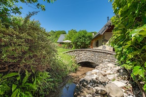 A stone bridge over a stream, surrounded by greenery and a house with a sloped roof.