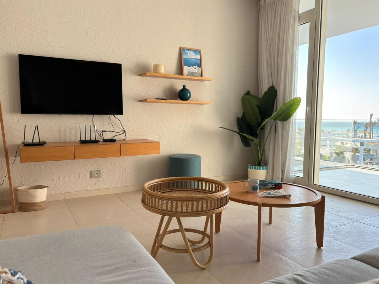 A modern living room with a flat-screen TV, wooden shelves, a round wicker chair, a coffee table, and a large window with a view.