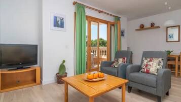 Sala de estar con televisor, mesa de centro de madera, dos sillones, un cactus y puerta corrediza de vidrio que da a un espacio exterior.