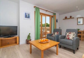 Sala de estar con televisor, mesa de centro de madera, dos sillones, un cactus y puerta corrediza de vidrio que da a un espacio exterior.