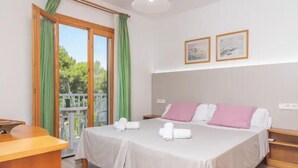 A hotel room with a bed, a desk, a balcony with curtains, and two framed pictures on the wall. - Apartment Son Baulo Poma (Santa Margalida)