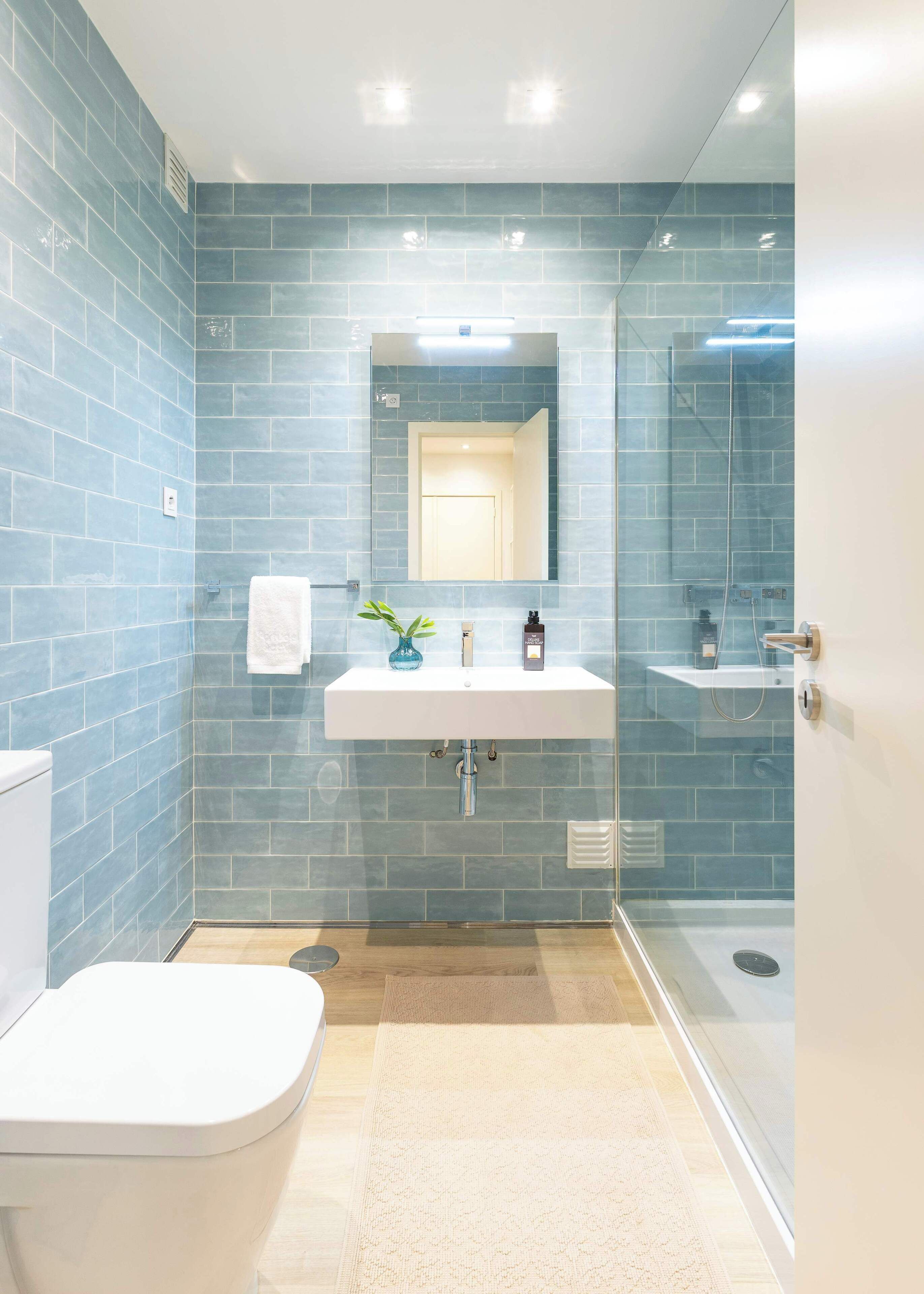 Ein Badezimmer mit weißem Waschbecken, Spiegel und Toilette, mit blauen Fliesen und einer Duschkabine aus Glas.