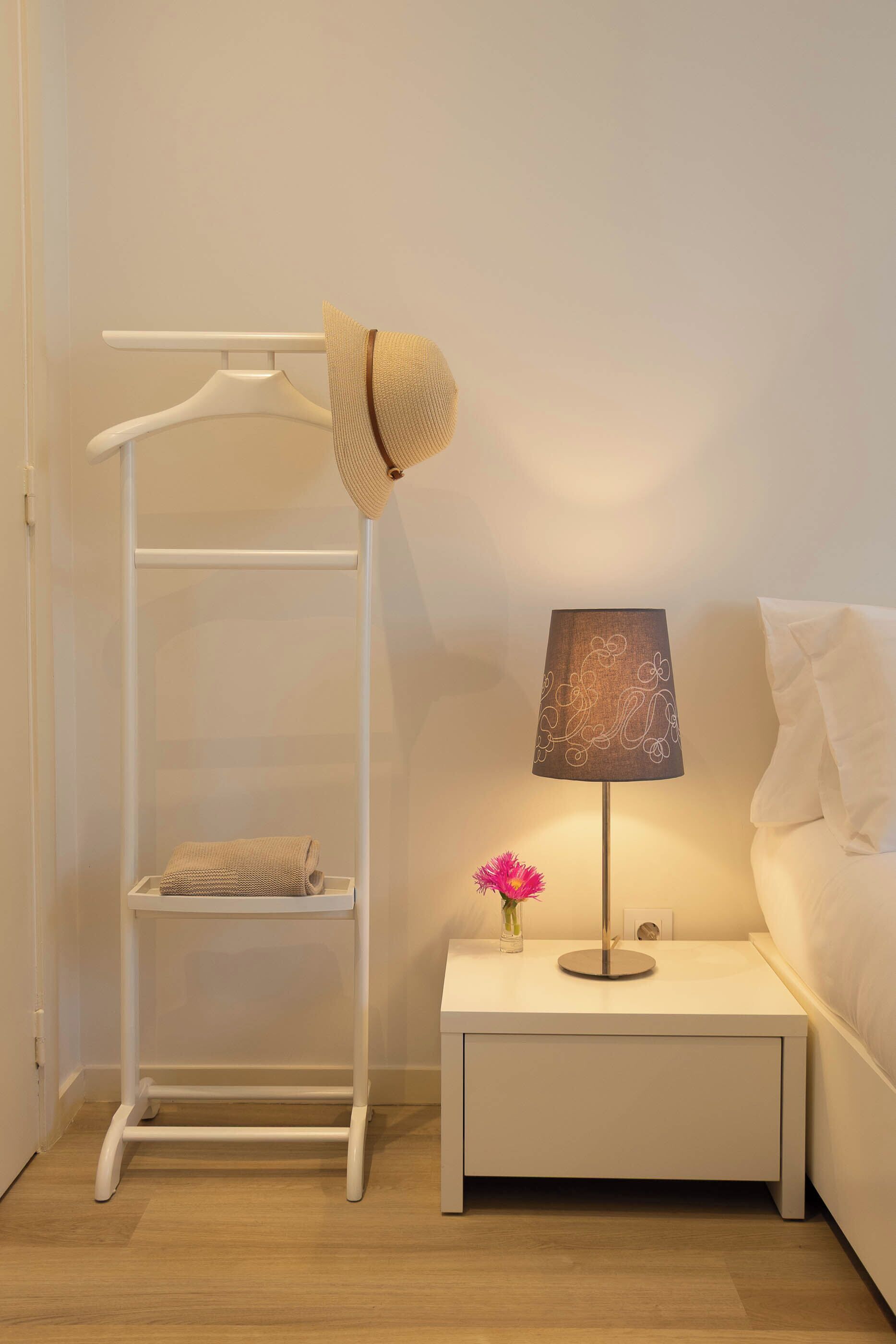 A white bedside table with a lamp, a white nightstand, a white wardrobe, and a hat on the wardrobe.