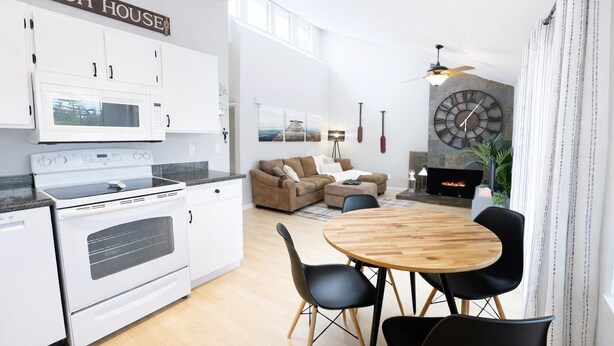 A modern kitchen with white cabinets, a microwave, and an oven. There is a dining area with a wooden table and black chairs. A sofa and a television are visible in the living area.