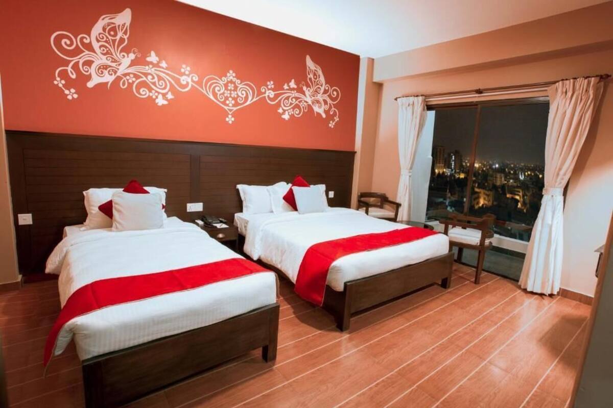 A hotel room with two beds, a nightstand, a chair, a window with curtains, and a city view.