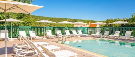 Un área al lado de la piscina con sillones de descanso y sombrillas, rodeada por una terraza de madera y una valla verde.