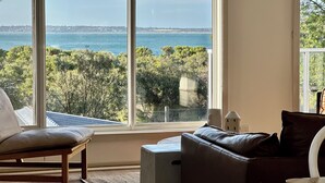 A room with a large window offering a view of the sea and trees. There is a brown leather sofa and a wooden chair. - On the beach, Cowes, Phillip Island, Sleeps (Cowes)