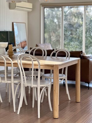 A dining area with a wooden table and white chairs, a brown sofa, and a large window with a view of trees. - On the beach, Cowes, Phillip Island, Sleeps (Cowes)