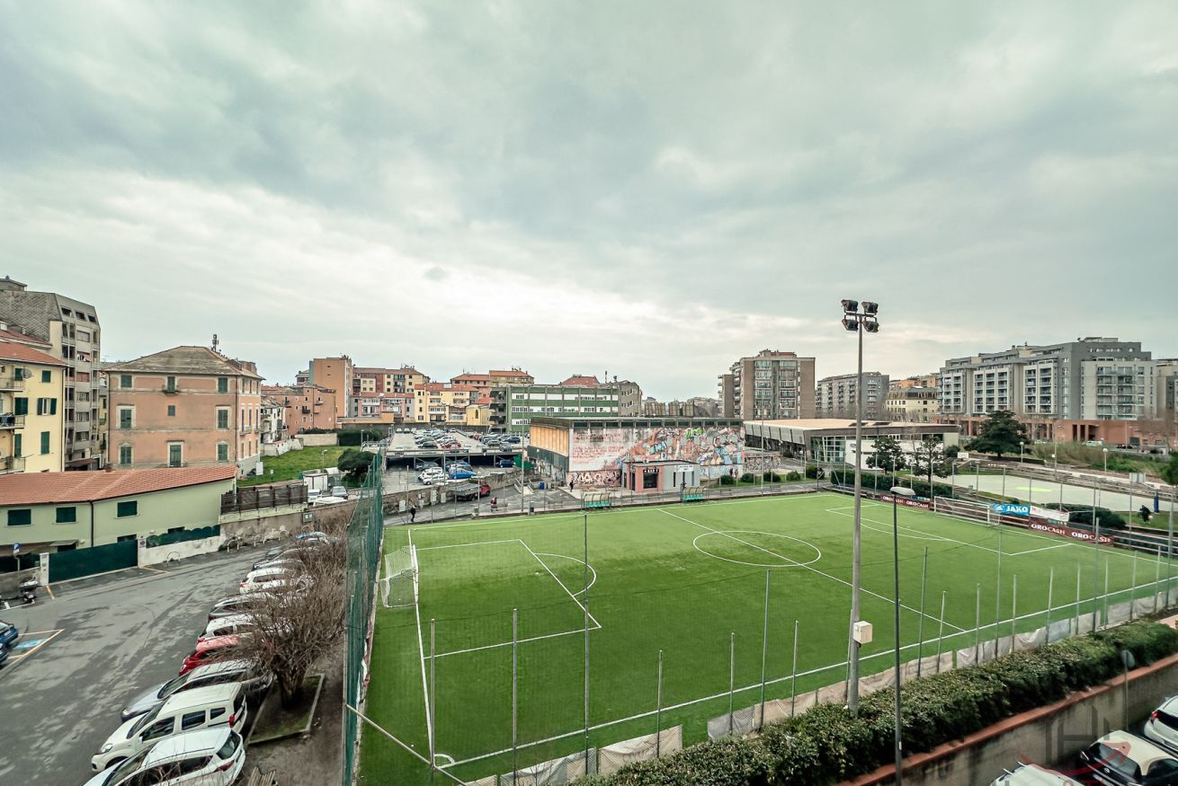 A soccer field surrounded by buildings, a parking lot, and a fence.