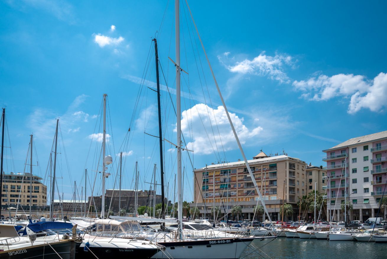 Una marina con diverse barche a vela ormeggiate, un edificio con balconi e un cielo azzurro limpido.