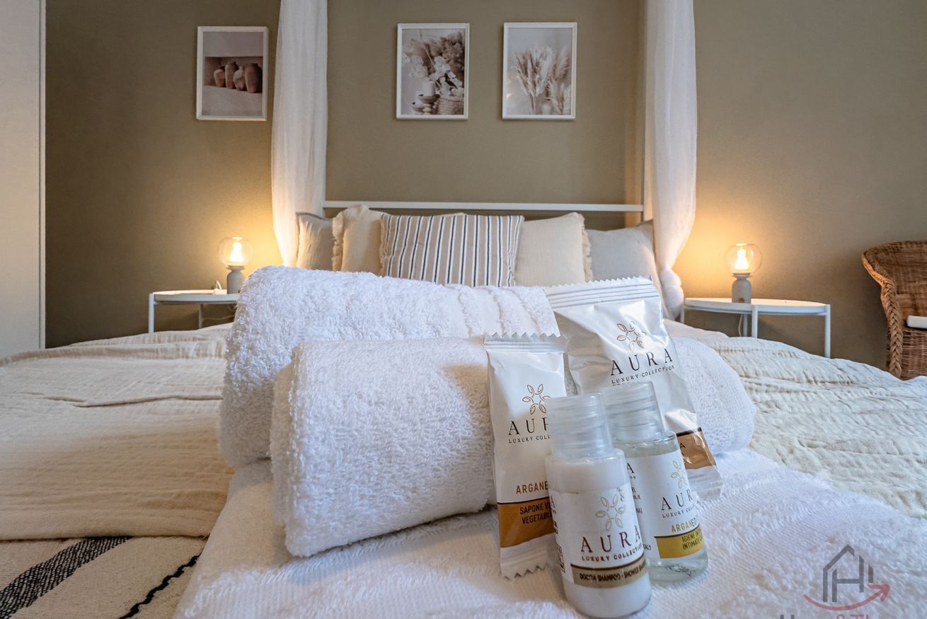 Uma cama bem arrumada com lençóis brancos, uma mesa de cabeceira com abajur e um criado-mudo com luminária.