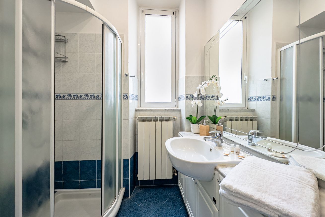 Ein Badezimmer mit Glasdusche, einem weißen Waschbecken, einem Spiegel und einer Topforchidee.