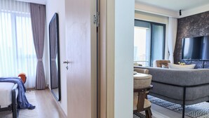 Quarto de hotel com cama, espelho, porta e vista para a sala de estar com sofá e televisão.