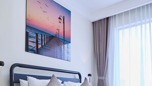 Quarto de hotel moderno com cama, mesa de cabeceira, pintura de um pôr do sol sobre um cais e janela com cortinas.