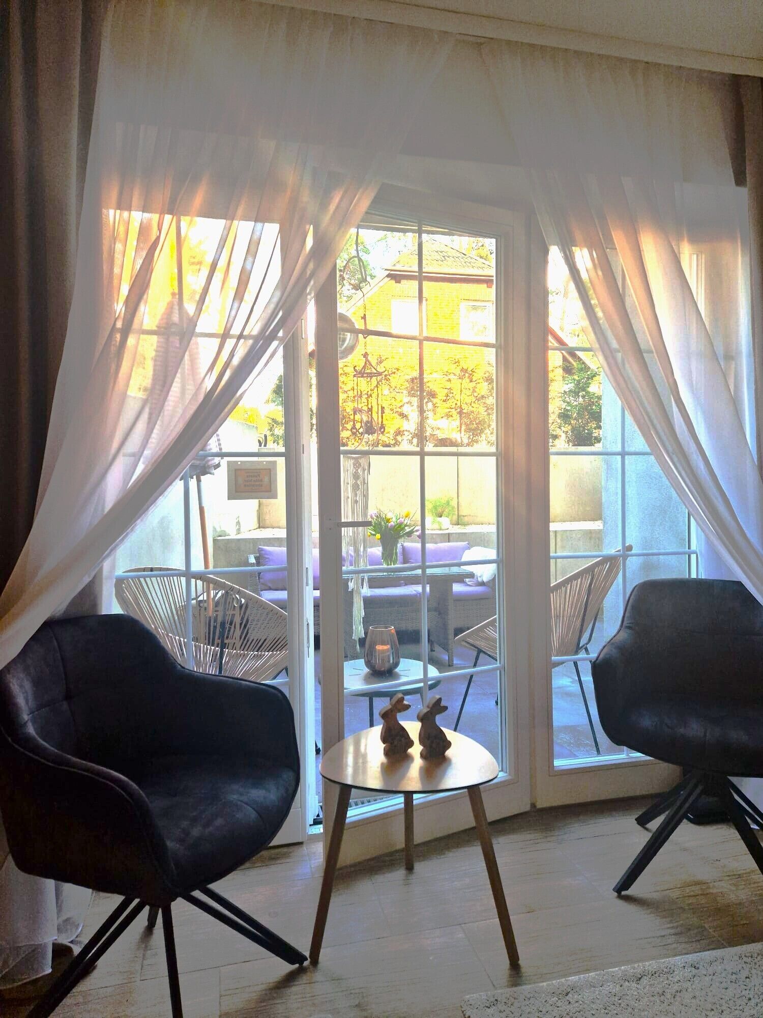 A room with two black chairs, a small round table, and a view through a glass door to an outdoor seating area with wicker furniture.