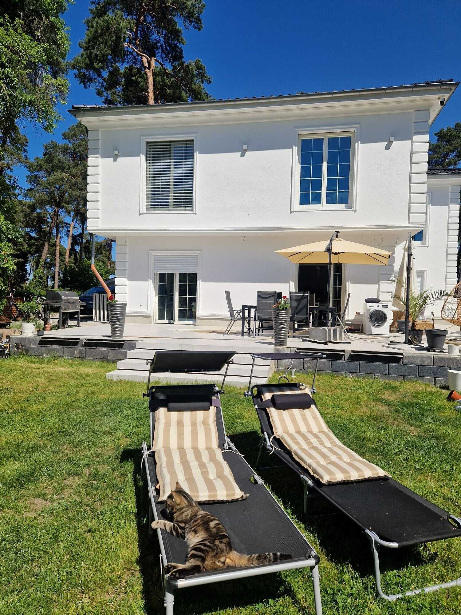 A two-story white house with a patio area featuring two lounge chairs and a table.