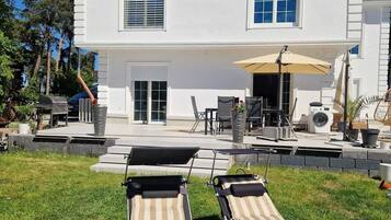 A two-story white house with a patio area featuring two lounge chairs and a table.