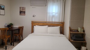Siististi järjestetty hotellihuone, jossa on sänky, työpöytä, tuoli, pieni hylly pyyhkeille ja seinälle asennettu ilmastointilaite.