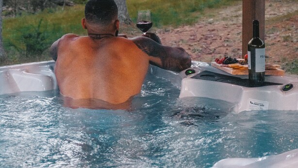 A person in a hot tub outdoors with a bottle of wine and a glass, surrounded by trees and a wooden structure. - Leisure Lake - 3BR Home, Hot Tub, Paddle boats (Scotrun)