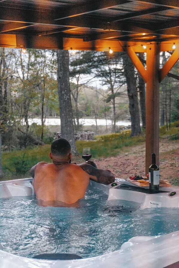 Eine Person entspannt sich in einem Außen-Whirlpool mit einer Weinflasche und einem Glas, umgeben von Bäumen und einer Holzkonstruktion.