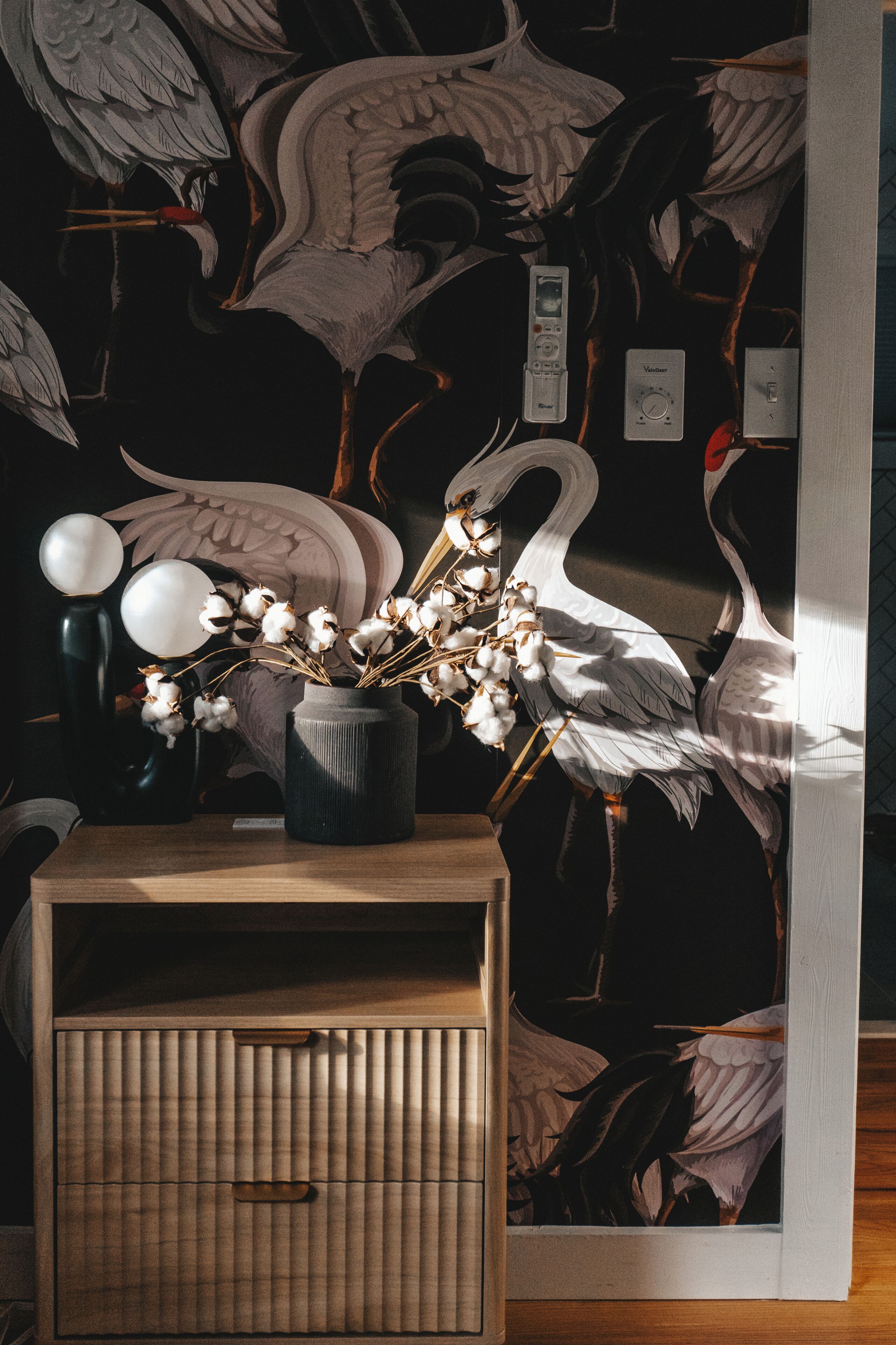 A room with a wall mural of birds, a wooden cabinet with a handle, and a vase with cotton plants.