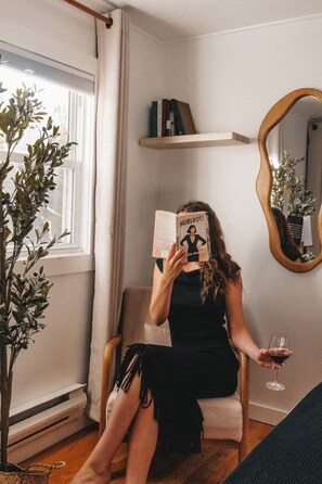 Una persona sentada en una silla leyendo un libro, con una planta y un espejo en la pared.