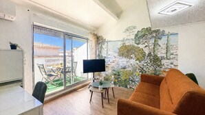 Ein Wohnzimmer mit einer Couch, einem Fernseher, einem Esstisch, Stühlen, einer Glastür zu einem Balkon und einem Wandgemälde.