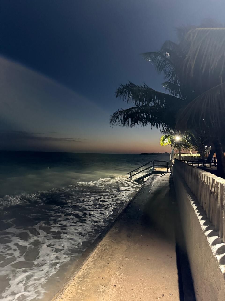 Uma praia à noite com calçadão de concreto, palmeiras e mar calmo.