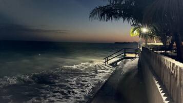 Uma praia à noite com calçadão de concreto, palmeiras e mar calmo.