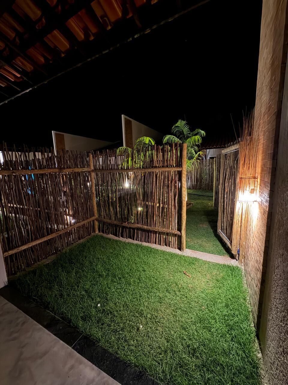 A nighttime outdoor area with a bamboo fence, artificial grass, and a pathway leading to a building.