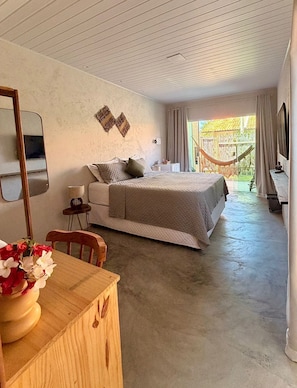 A bedroom with a bed, a desk with a chair, a mirror, and a view of a hammock outside.