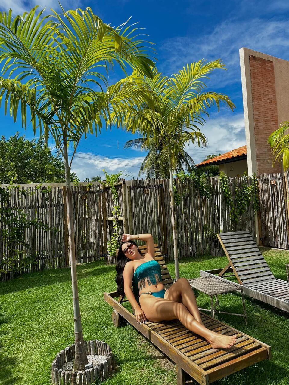 Una persona descansando en una terraza de madera en un patio trasero con valla de bambú, palmeras y una casa al fondo.