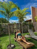 Una persona descansando en una terraza de madera en un patio trasero con valla de bambú, palmeras y una casa al fondo.