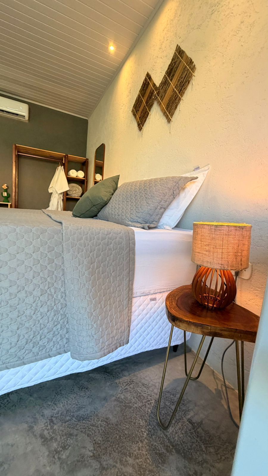A bedroom with a bed, pillows, a bedside lamp, a small table, and a wall-mounted mirror.