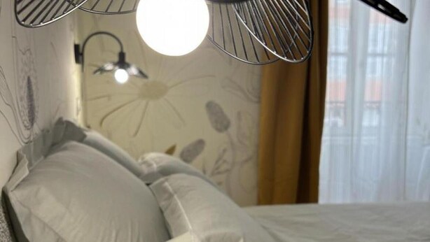 Una cama con ropa blanca, una lámpara de pared y un ventanal con cortinas.
