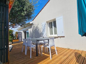 Una terraza de madera con una mesa y sillas, un muro exterior blanco y una ventana con contraventanas.