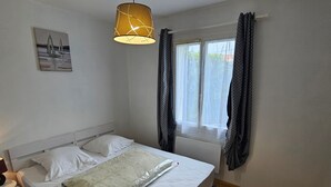 A bedroom with a bed, a bedside table, a lamp, a window with curtains, and a painting on the wall. - Single-storey house with quiet garden - 6 guests (Royan)