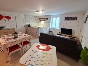 A living room with a sofa, a TV, a dining table with chairs, a calendar, and a kitchen in the background. - Charming cottage with parking - 2 guests in village center (Choloy-Ménillot)