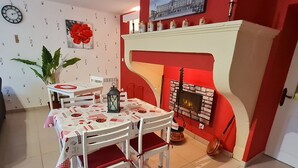 A dining area with a white table and chairs, a red and white patterned tablecloth, a large potted plant, a clock on the wall, and a fireplace with a red mantle. - Charming cottage with parking - 2 guests in village center (Choloy-Ménillot)