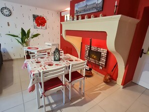 A dining area with a white table and chairs, a red and white patterned tablecloth, a large potted plant, a wall clock, and a fireplace with a red mantle. - Charming cottage with parking - 2 guests in village center (Choloy-Ménillot)