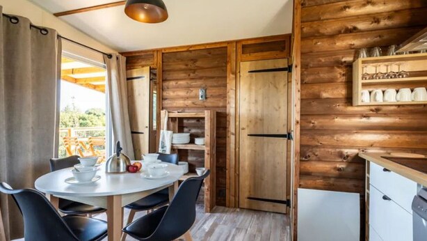A wooden interior with a dining table, chairs, a kitchen area, and a view through an open door.