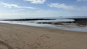 Uma praia com pegadas, céu claro e terra ao longe.