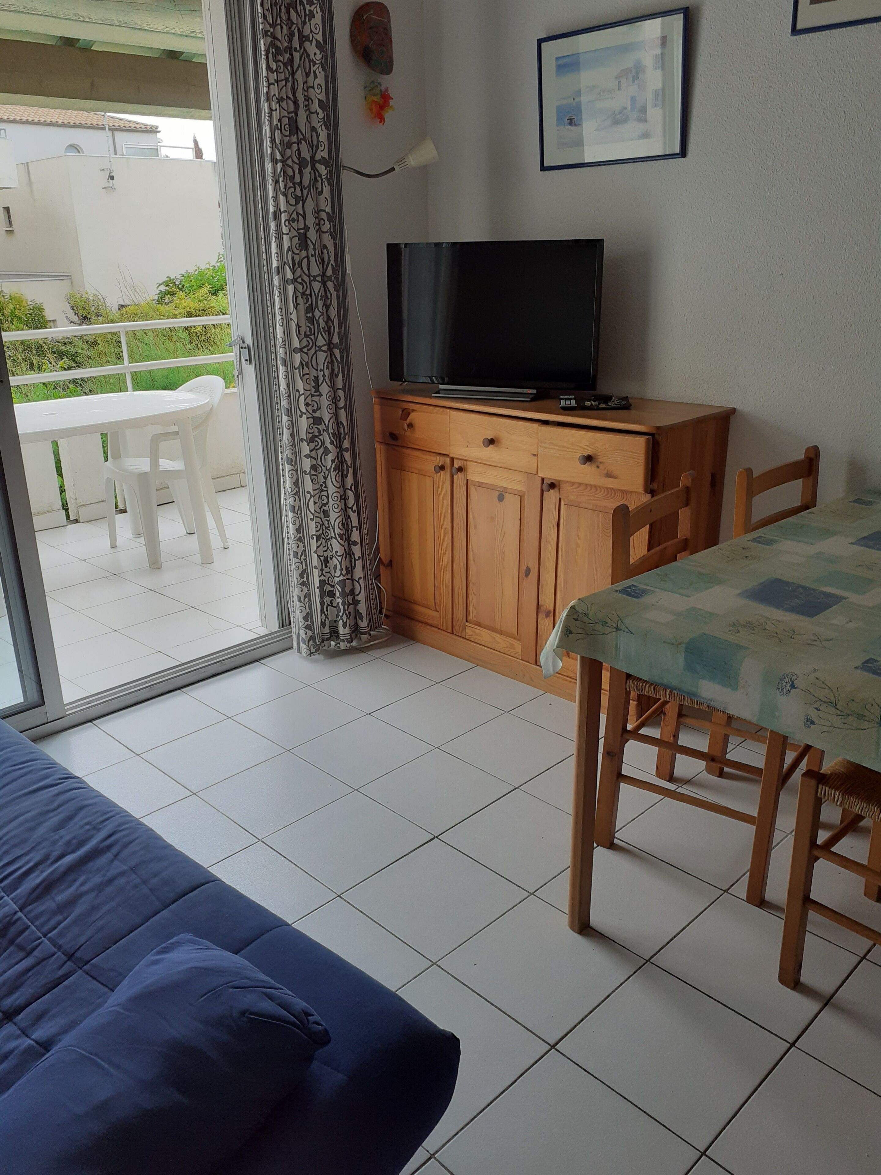 A room with a bed, a TV on a wooden cabinet, a dining table with chairs, and a view through a glass door.