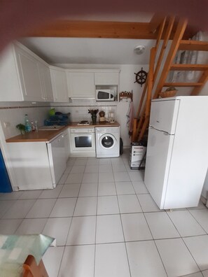 A compact kitchen with white cabinets, a microwave, a washing machine, and a refrigerator. - AGREABLE t2 mezzanine- SEASIDE (LE GRAU DU ROI)